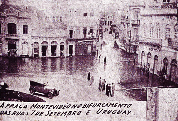 foto de um largo semi-tomado pelas águas, com algumas pessoas e um carro estacionado, e uma esquina alagada ao fundo com a legenda “A praça Montevideo no bifurcamento das ruas 7 de setembro e Uruguay”.